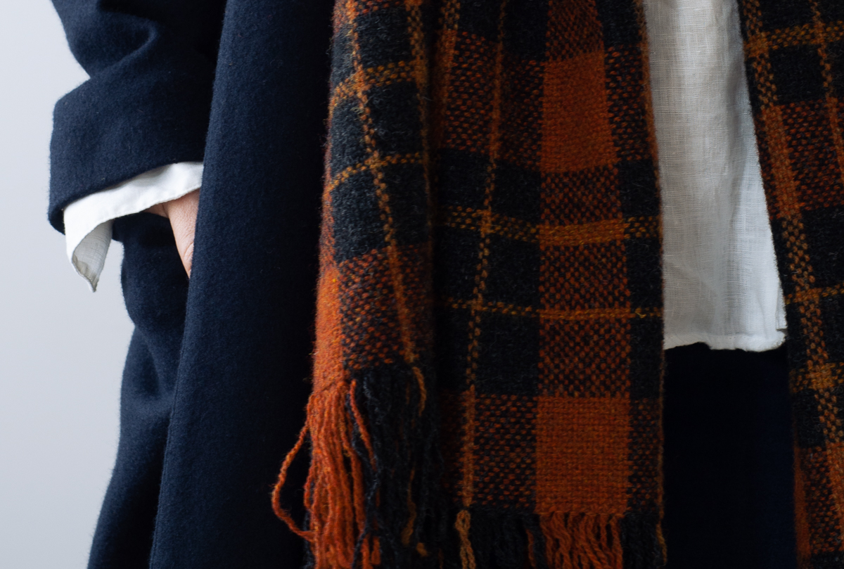 Handwoven check scarf in shades of burnt red, coal and terracotta worn over a navy wool coat and crisp white linen shirt.