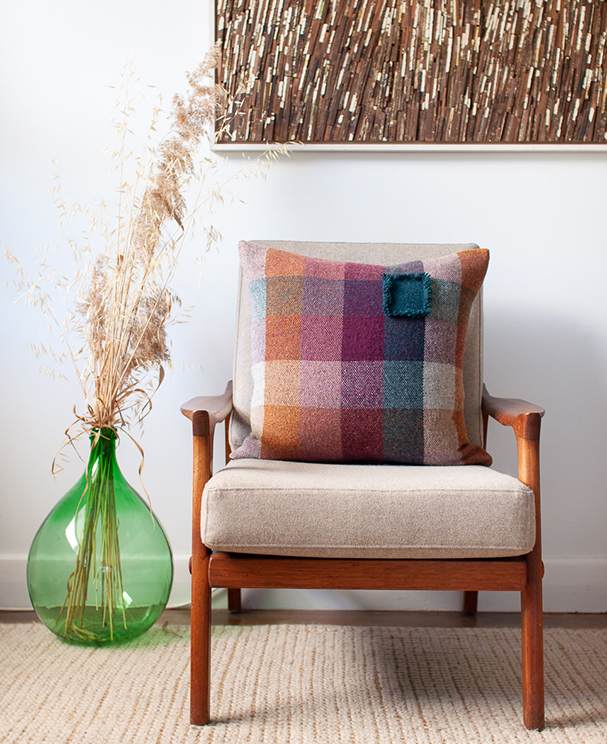 5 colour check cushion in warm shades with a short teal blue fringe on one check resting on a Danish Deluxe chair next to a green vintage demijohn.
